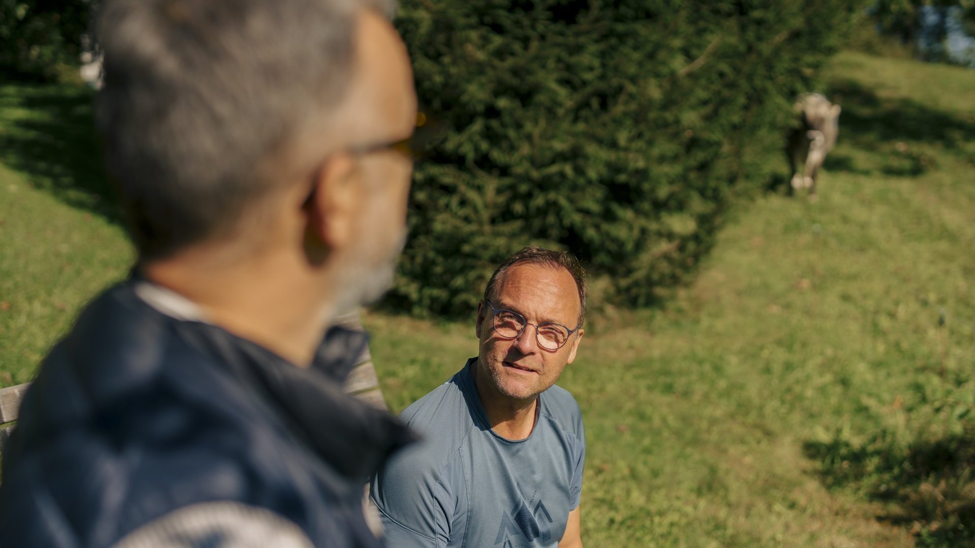 Zwei Herren unterhalten sich, einer sitzt auf einer Holzbank, der andere steht daneben.
