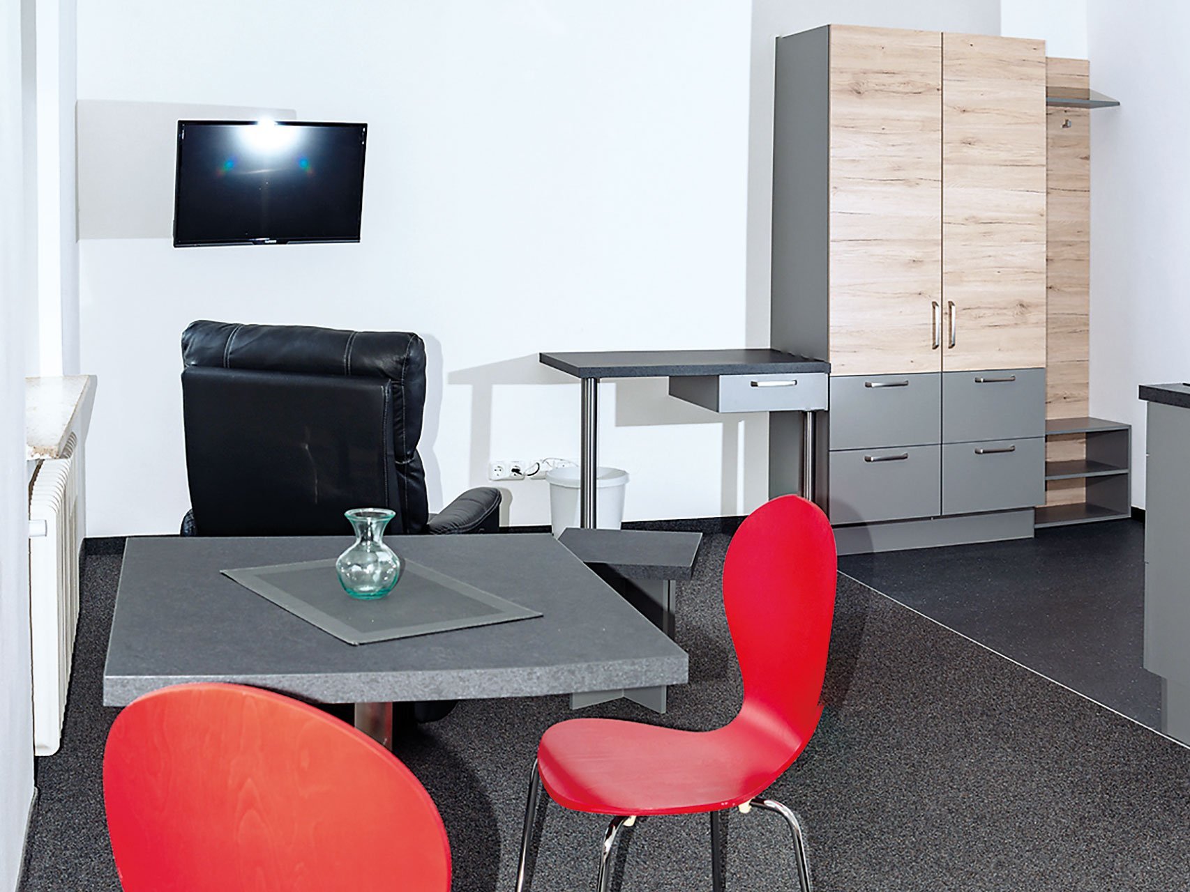 Ein funktionaler, moderner Raum mit Fernseher, einem Schrank, Ledersessel und einem Tisch mit roten Stühlen