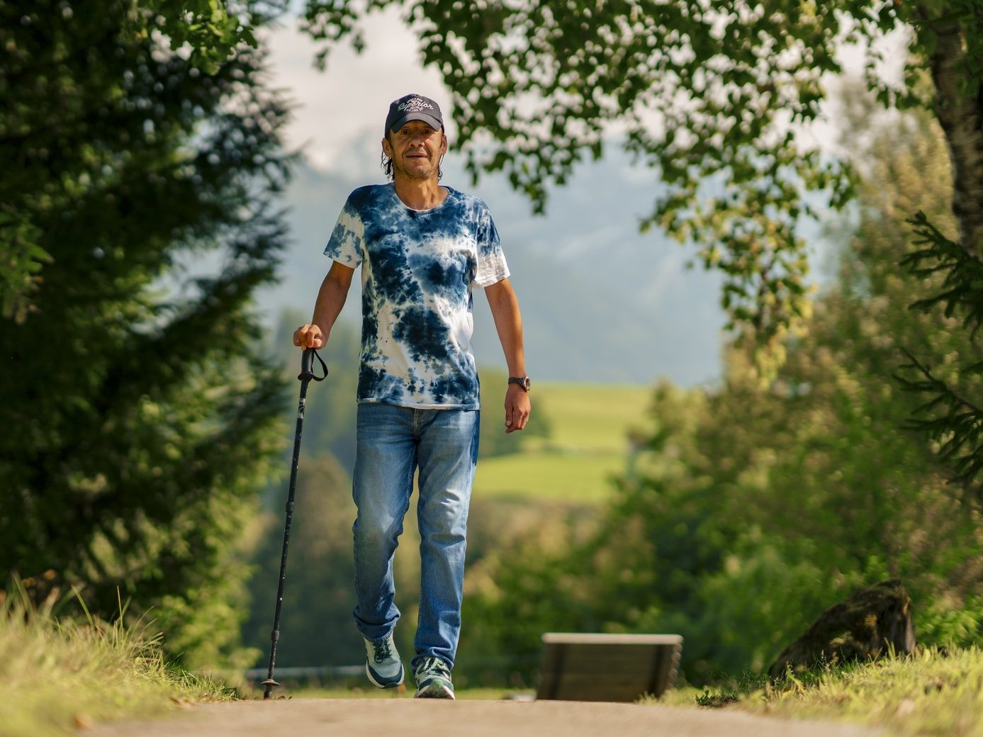 Ein Mann macht Nordic Walking auf einem Weg durch den Wald. Er trägt ein blau-weißes T-Shirt, eine Jeans und eine Kappe.