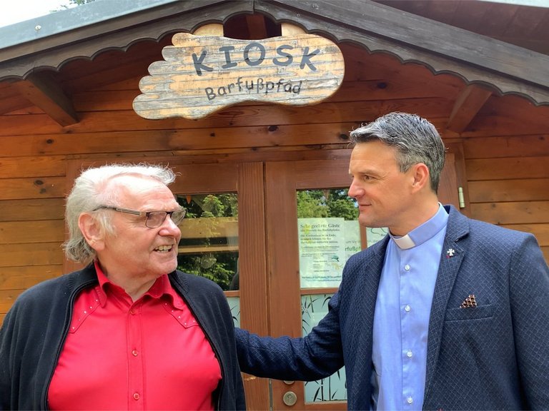 Pater Jörg vor dem Kiosk "Barfußpfad" hört einem älteren Mann zu