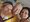 Drei lächelnde Personen posieren eng beieinander für ein Foto. Links eine Frau mit blonden Haaren und Namensschild, in der Mitte ein älterer Mann im gelben Hemd und rechts eine Frau mit dunklen Haaren in einem Beige-Blazer. Im Hintergrund ist ein Gemälde an der Wand zu sehen.