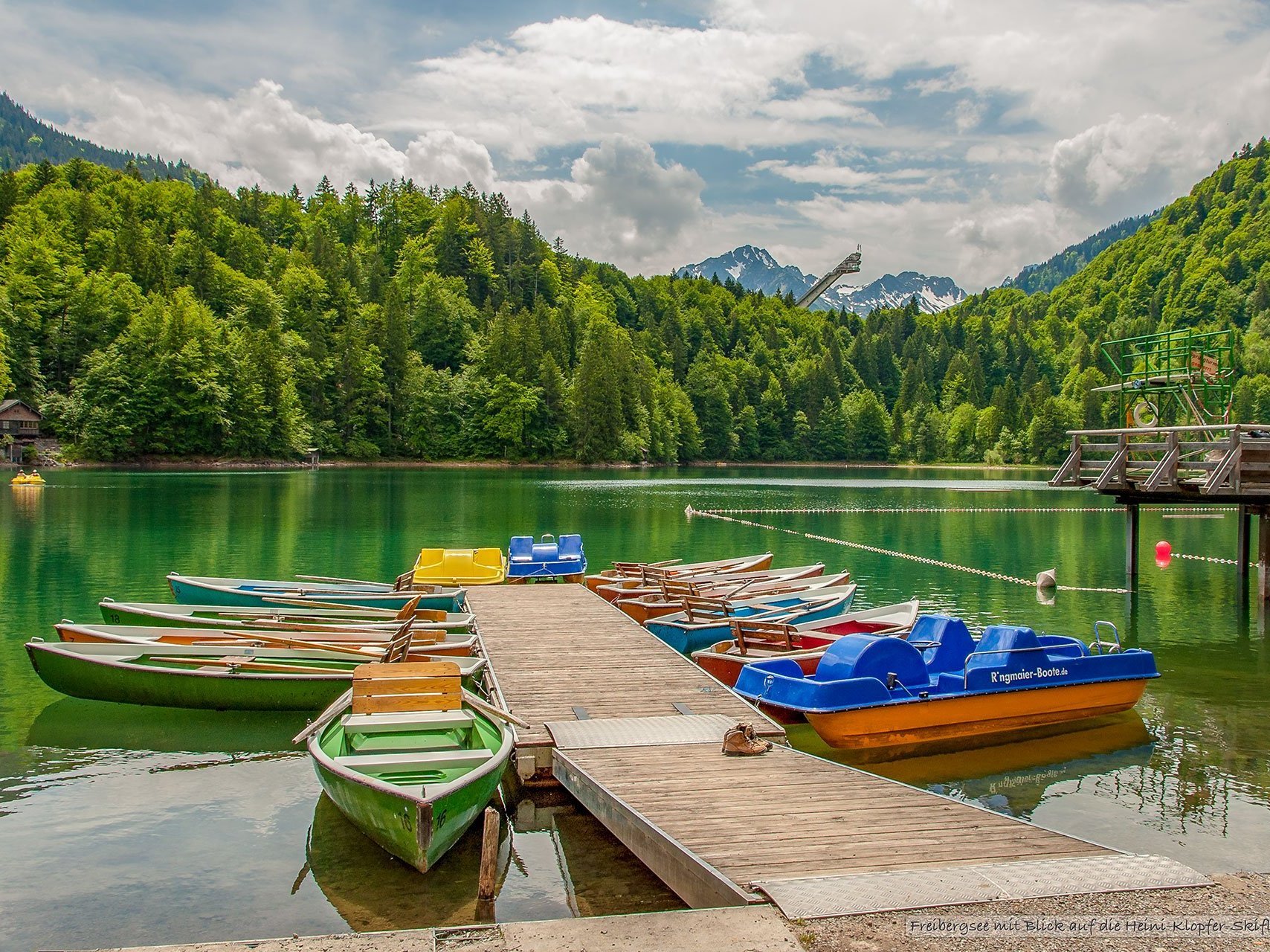 Badesee mit Ruderbooten am Steeg - im Hintergrund eine Skisprungschanze und die Alpen